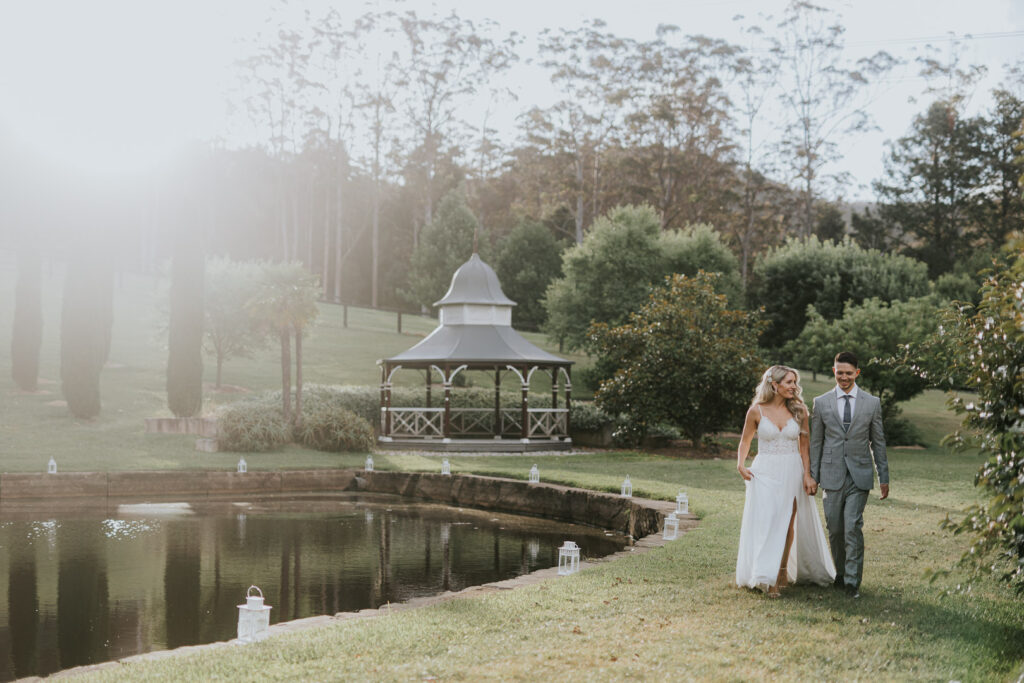 Fernbank Farm, Yarramalong on the NSW Central Coast. The perfect place for your Country Elopement. A couple walks through the grounds by a lake with a pretty band stand behind the 