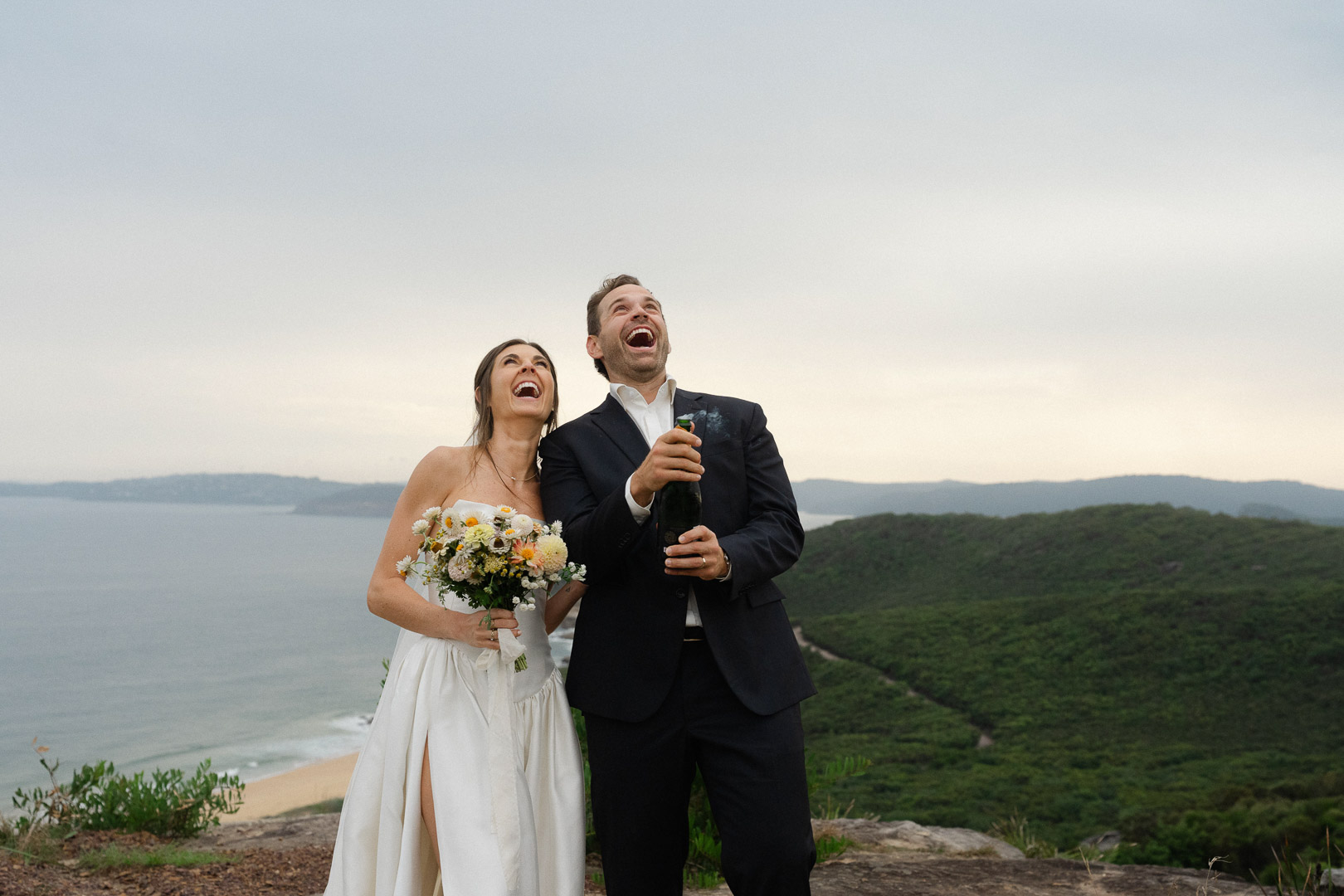 How to plan a Central Coast NSW Elopement: Man and woman popping a bottle of champagne with a view of the Hawkesbury River and coastline behind them